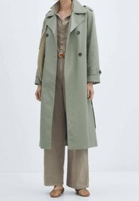 Manteau trench vert clair avec des boutons double boutonnage, larges revers et une silhouette longue, superposé sur une combinaison khaki et un pantalon large.
