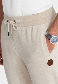 Beige trainingsbroek met een trekkoord in de taille, gestructureerde stof, zijzakken en een hexagonaal bruin merklabel op de dij.