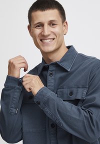 Jeans jakna v temno modri barvi, z zapiranjem na gumbe, dvema žepoma na prsih, z gumbki na manšetah in sproščenim krojem. Gladka tekstura, standarden ovratnik.