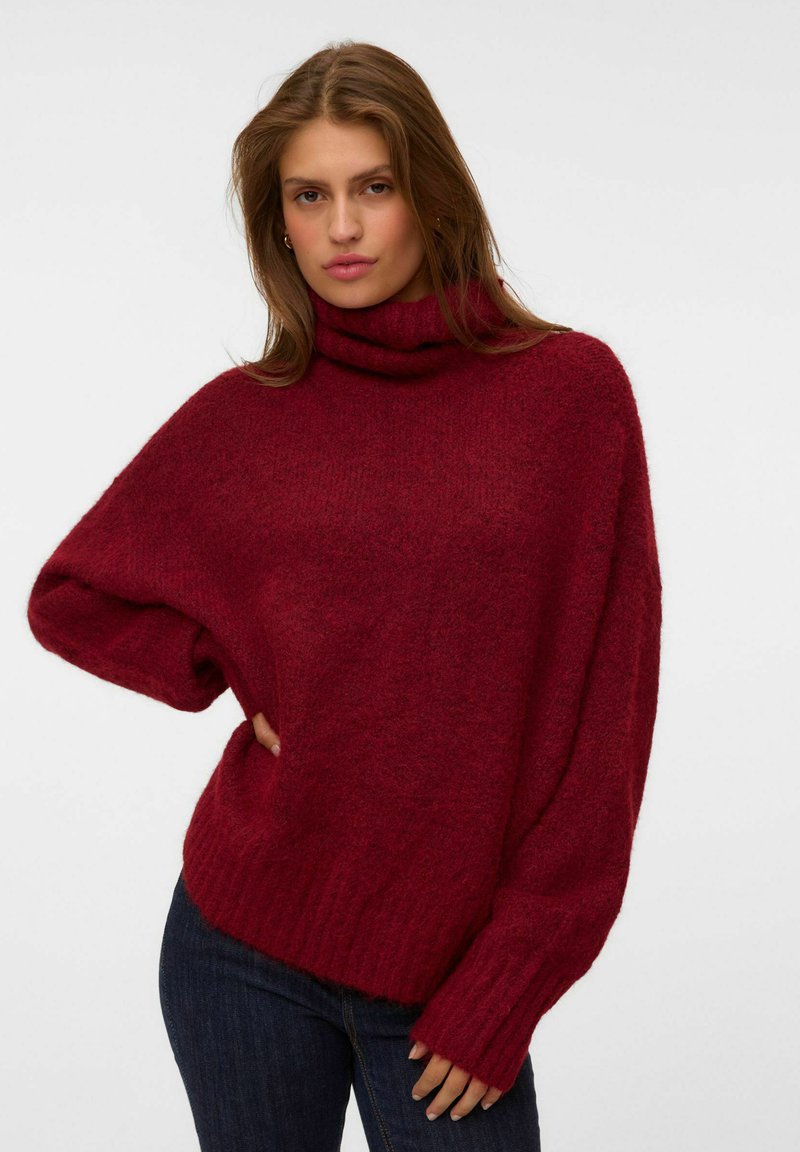 Jeune femme aux longs cheveux portant un pull à col roulé bordeaux ample et duveteux ainsi qu'un jean foncé, debout devant un fond clair uni.