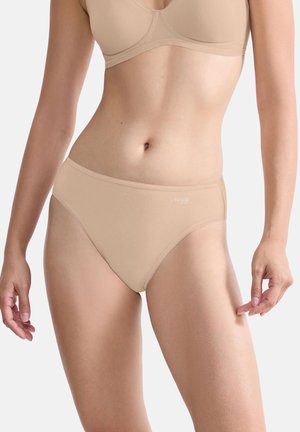 Frau trägt nahtlosen beigen BH und passende hochgeschnittene Slips, steht vor neutralem Hintergrund.