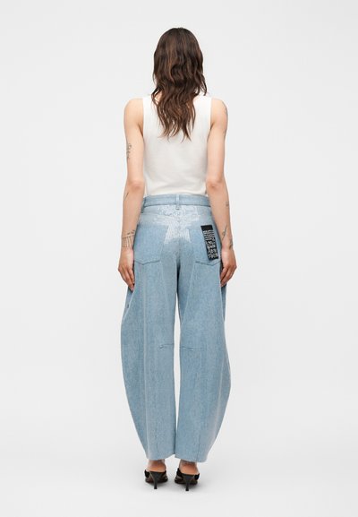 Γυναίκα με κυματιστά μαλλιά, που φοράει αμάνικη λευκή μπλούζα, φαρδιά ανοιχτό μπλε τζιν με σχέδιο αστεριών και μαύρο μπαλώμα στην πίσω τσέπη, μαύρες γόβες.