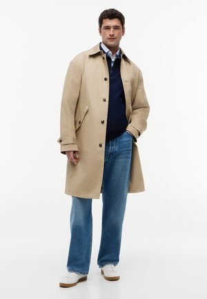Mann trägt beige Trenchcoat, navy Pullover, blaue Jeans und weiße Sneakers, steht mit einer Hand in der Tasche vor weißem Hintergrund.