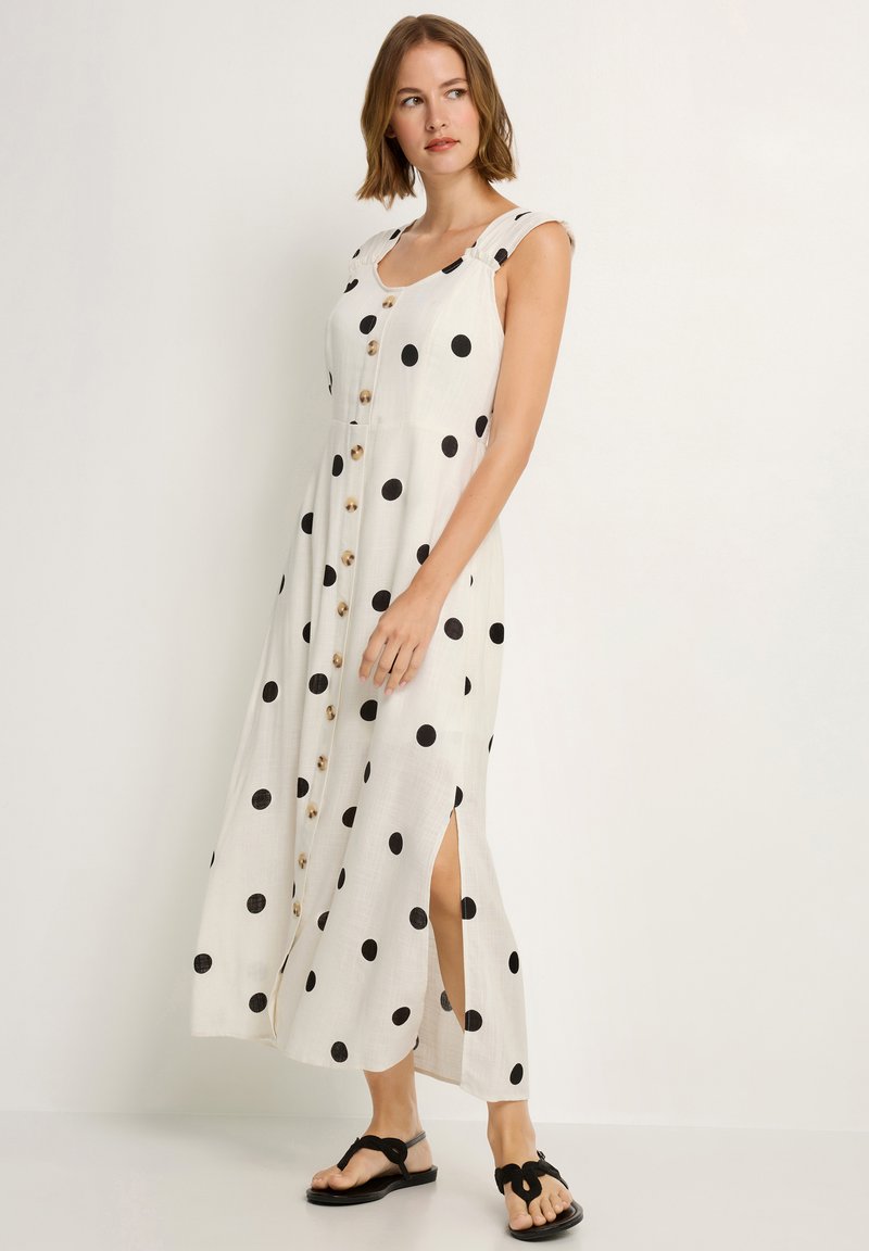 Weißes Kleid mit schwarzen Punkten, knielang, mit Knöpfen vorne und seitlichen Schlitzen. Kombiniert mit schwarzen Sandalen.