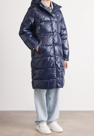 Personne portant un long manteau matelassé bleu marine avec une capuche, un jean bleu clair et des baskets blanches, debout sur un sol gris contre un mur blanc.