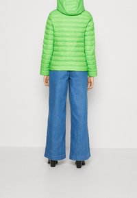 Veste matelassée verte avec capuche, présentant une texture matelassée et une taille ajustée. Associée à un pantalon en denim à jambes larges et des bottes noires à talons.
