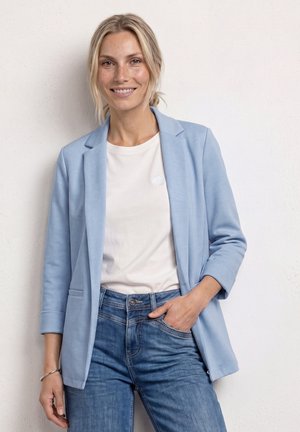 Femme souriante aux cheveux blonds portant un blazer bleu clair, un t-shirt blanc et un jean bleu, debout devant un mur blanc uni.