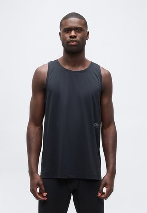 Atletski moški s temno poltjo, oblečen v brezrokavnik črno športno majico in črne kompresijske pajkice, stoji pred belim enobarvnim ozadjem.