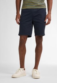 Námořnické modré kraťasy vyrobené z bavlny, se standardním střihem, dvěma kapsami a čistým provedením. Nosí se s krémovými teniskami.