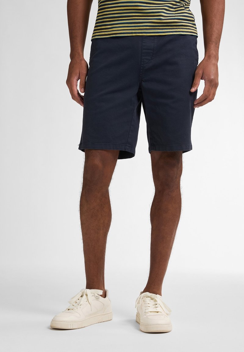 Námořnické modré kraťasy vyrobené z bavlny, se standardním střihem, dvěma kapsami a čistým provedením. Nosí se s krémovými teniskami.