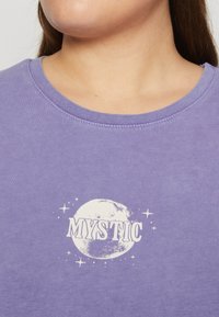 Purple crewneck shirt featuring a white graphic of a moon and the word "MYSTIC" below it, with small star accents on a soft texture.