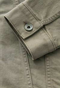 Tela de algodón de color verde oliva claro con un puño doblado que presenta un botón a presión de metal grabado con "DENIM SUPPLY CO." y una costura cuidada.