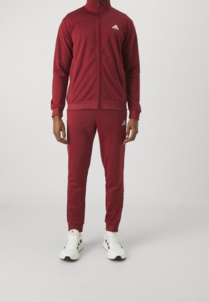 Mann trägt einen roten Adidas Trainingsanzug mit durchgehendem Reißverschluss, passenden Hosen und weißen Adidas Sneakers, steht vor einem schlichten Hintergrund.