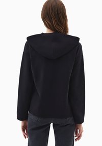 Černá mikina s volným střihem, vybavená velkou kapucí, dlouhými rukávy a hladkou texturou. Nosit s tmavými džínami.