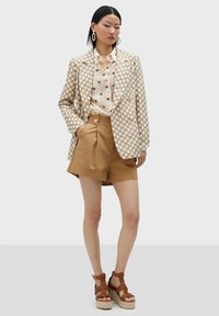 Beige mönstrad blazer över en vit skjorta med bruna prickar, tillsammans med bruna plisserade shorts och bruna remsandaler med kilklack.