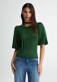 Top verde texturizzato con maniche a sbuffo, caratterizzato da un collo legato. Abbinato a jeans in denim blu, che mostrano una silhouette aderente.