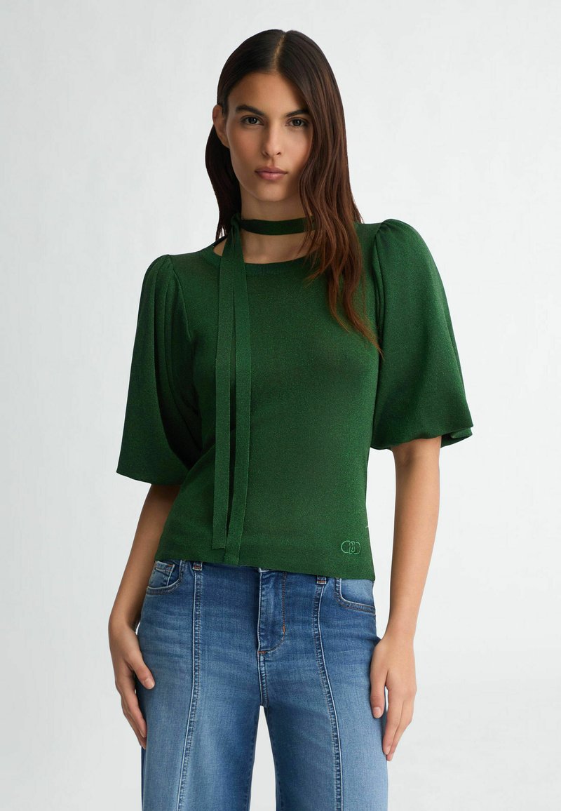 Top verde texturizzato con maniche a sbuffo, caratterizzato da un collo legato. Abbinato a jeans in denim blu, che mostrano una silhouette aderente.