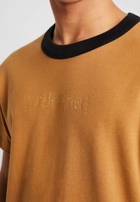Detailní záběr na osobu, která má na sobě hnědé tričko s černým límečkem a na hrudi má vyšitý nápis "undefined".