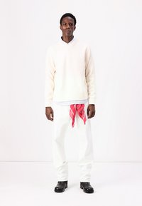 Un pull crème avec un col montant, une chemise blanche en dessous, un pantalon blanc et un bandana rose noué à la taille. Des bottes noires complètent le look.