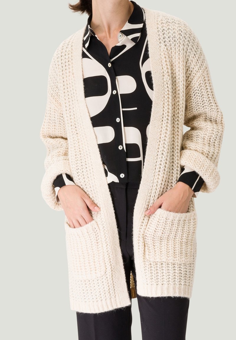 Femme portant un cardigan en grosse maille crème avec poches avant, sur une chemise à boutons noir et blanc à motifs, et un pantalon noir.