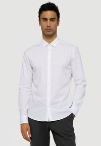 Bílá košile s dlouhým rukávem, špičatým límečkem, zapínáním na knoflíky a slim fit střihem. Vyrobena z hladkého materiálu, s diskrétním logem na hrudi.