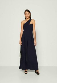 Navy blue, one-shoulder jumpsuit made from a smooth fabric; wide-leg design with pleats; accessorized with a small bag and high-heeled shoes.