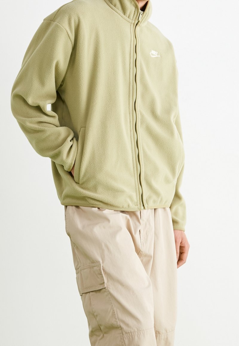 Ljusgrön fleece zip-up jacka med hög krage och sidfickor, bärs över beige cargobyxor. Har en vit logotyp på bröstet.