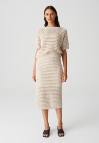 Beige gestricktes Zweiteiler-Outfit, bestehend aus einem kastigen Kurzarm-Oberteil und einem figurbetonten Midi-Rock mit Rautenmuster. Getragen mit schwarzen Schuhen.