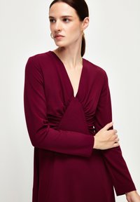 Burgundivioletti mekko pitkillä hihoilla, V-pääkohdalla ja rypytetty yksityiskohta rinnan kohdalla, valmistettu sileästä kankaasta, luoden vartalonmyötäisen siluetin.