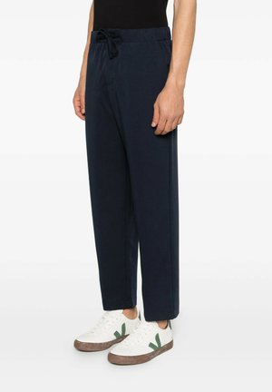 Uomo con pantaloni blu navy con coulisse e sneakers bianche con dettagli verdi, in piedi su uno sfondo chiaro uniforme.