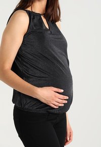 Svart ärmlös gravidblus med en glänsande yta, nyckelhålsutskärning och draperad nederkant, bärs över svarta byxor.