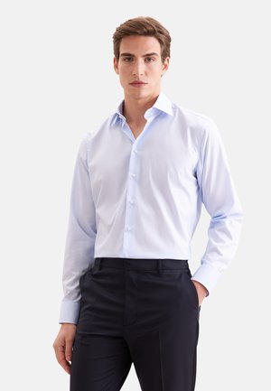 Homme portant une chemise bleu clair à manches longues boutonnée et un pantalon noir, debout avec une main dans la poche sur un fond uni.