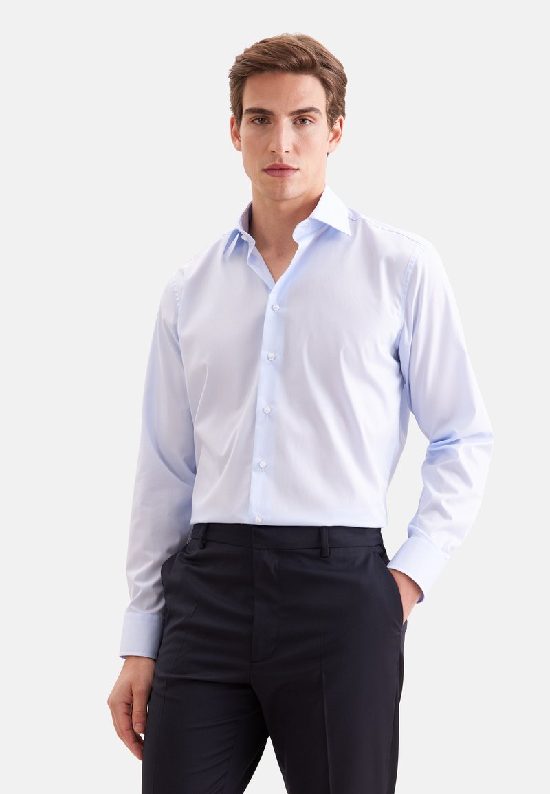 Homme portant une chemise bleu clair à manches longues boutonnée et un pantalon noir, debout avec une main dans la poche sur un fond uni.