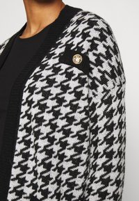 Gebreide cardigan met een zwart-wit hounds-tooth patroon, een zwarte kraag en een gouden embleem op de borstzak.