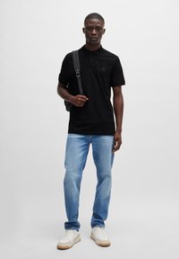 Schwarzes Polohemd, blaue Jeans, weiße Sneaker. Modell hält einen schwarzen Rucksack. Einfaches Design, einfarbige Farben, lässige Passform.
