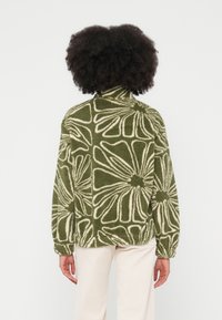 Sudadera verde de forro polar con estampado floral blanco, cuello alto y puños ajustados. Modelo visto desde atrás, vistiendo pantalones color crema.