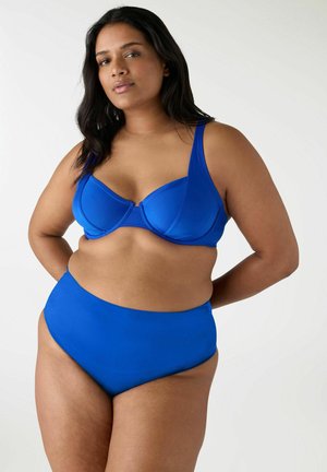 Donna con lunghi capelli scuri che indossa un reggiseno blu con ferretto e slip bikini a vita alta, in piedi con le braccia dietro la schiena.