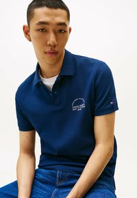 Navyblauwe poloshirt met korte mouwen, voorzien van een kraag en geborduurd logo op de borst. Gecombineerd met blauwe denim jeans.