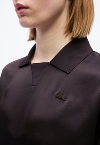 Femme aux cheveux blonds courts portant une chemise noire à col avec un petit logo crocodile noir brodé sur la poitrine.