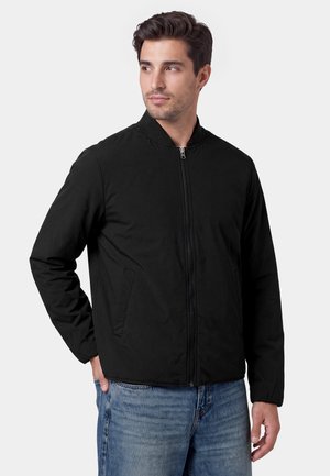 Mann trägt eine schwarze Jacke mit Reißverschluss und blaue Jeans, steht mit einer Hand in der Tasche und schaut seitlich vor neutralem Hintergrund.
