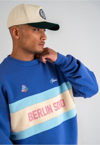 Sweatshirt albastru cu o banda orizontala bej și albastru deschis, având inscripția "BERLIN SOHO" în roz. Material din bumbac, cu guler rotund și un detaliu mic de logo. Şapcă crem.