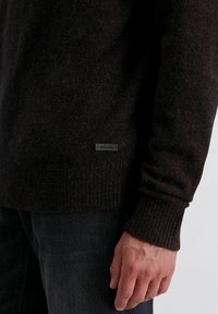 Rechter Arm und Hand einer Person, die einen dunkelbraunen Strickpullover mit geripptem Bündchen und Saum sowie dunklen Jeans trägt, vor einem weißen Hintergrund.