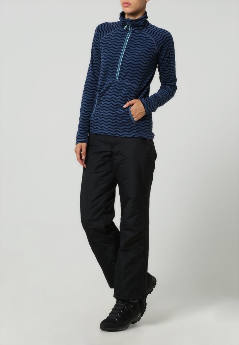 Femme portant une polaire bleue à motif zigzag avec une demi-fermeture éclair et un pantalon de plein air noir, debout avec une main dans la poche.