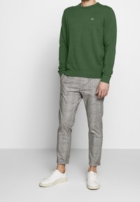 Maglione verde a maglia con collo tondo, abbinato a pantaloni slim-fit grigi a quadri e sneakers bianche. Caratteristiche includono polsini e orlo a coste.