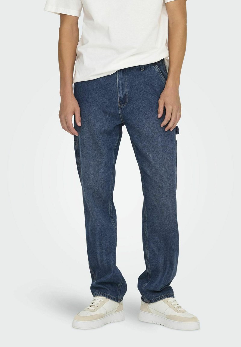 Jean en denim bleu foncé avec une coupe décontractée, arborant un design classique à cinq poches et des coutures discrètes. Associé à des baskets blanches.