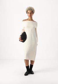 Λευκό πλεκτό φόρεμα με ανοιχτούς ώμους, μακριά μανίκια, μεσαίου μήκους σχέδιο, συνδυασμένο με μαύρο υφασμάτινο τσαντάκι και μαύρες κάλτσες με ραβδωτές άκρες.