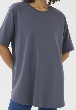 Person iført en løs, kortærmet, mørkegrå T-shirt med rund hals og hænderne bag ryggen mod en ensfarvet baggrund.