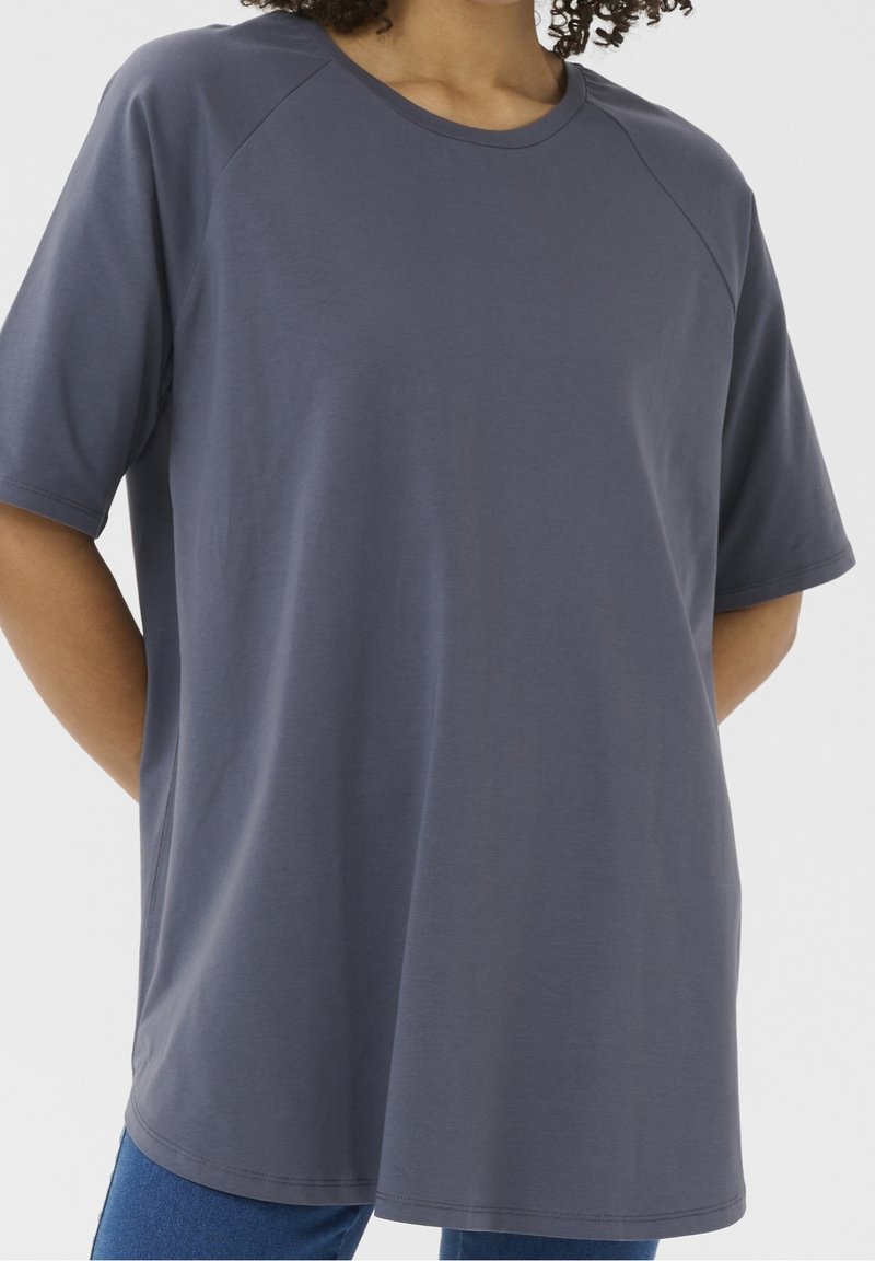 Personne portant un t-shirt ample à manches courtes, col rond, gris foncé, les mains dans le dos, sur un fond uni.