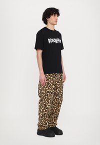 Melnas kokvilnas T-kreklam ar baltu grafiku; brīva piegriezuma brūnas leoparda prints bikses; melni slip-on apavi. Modelis stāv profila attēlā.