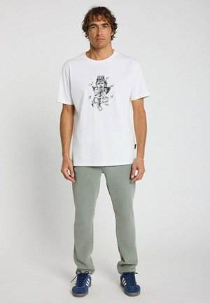 T-shirt blanc en coton avec un imprimé graphique noir représentant une figure angélique. Le modèle porte un pantalon côtelé vert clair et des baskets bleues.